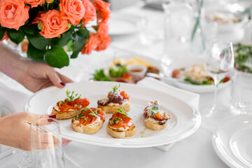 Hand holding a plate of assorted bruschettas on a festive table with glassware and a bouquet of orange roses, showcasing elegant food presentation and dining atmosphere.