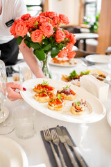 Waiter serving bruschetta at a restaurant table set with cutlery, wine glasses, napkins, and a vibrant bouquet of orange roses, capturing elegant dining atmosphere indoors.