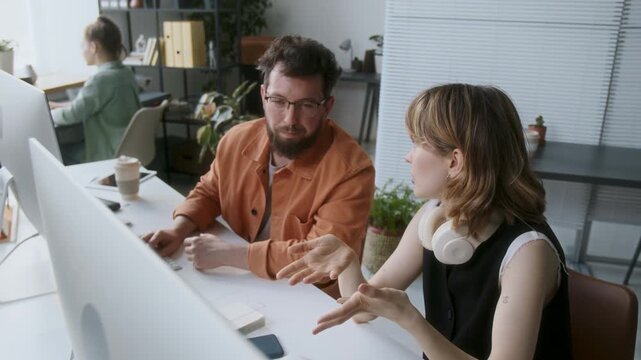 High angle shot of two male and female programmers sitting at wooden desk with two computers and office supplies while discussing new app glancing at monitor occasionally