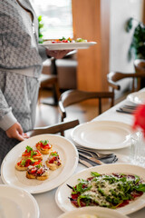 Person serving bruschetta with tomatoes and greens at a well-set table during a casual indoor meal or gathering.