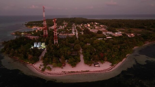 Aerial view of communication towers piercing the skyline above lush green trees on an island with a white beach at sunset, Goidhoo, Baa Atoll, Maldives.