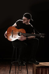 A man sits on a stool wearing a black outfit and sunglasses, playing an acoustic guitar. The dim...