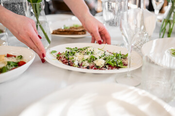 Top view of carpaccio salad with arugula and cheese being served at an elegant dining table with flowers, glassware, and cutlery in a fine restaurant setting.