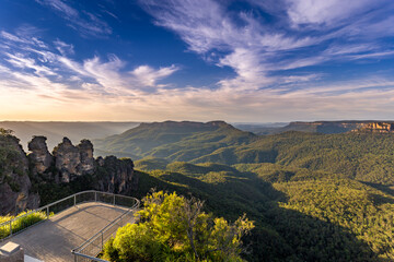 A panoramic view of the Three Sisters rock formation in the Blue Mountains, Australia. The photo was taken from the scenic Echo Point Lookout with a railing overlooking the vast landscape.