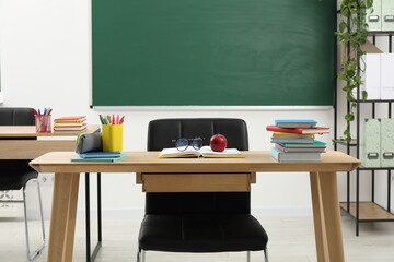 Different school stationery, glasses and red apple on wooden table near blackboard indoors