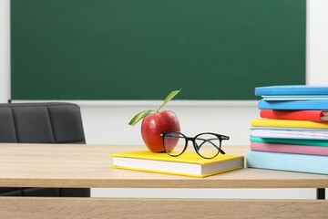 Different books, glasses and red apple on wooden table near blackboard indoors, space for text