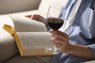 Woman with glass of wine reading book on sofa at home, closeup