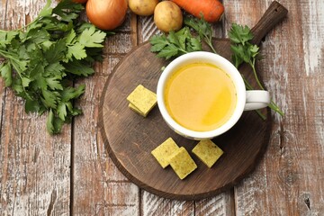 Aromatic bouillon cubes, broth, parsley and vegetables on wooden table, flat lay