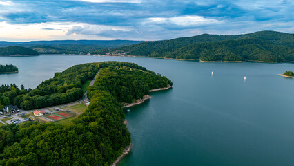 Obraz premium Aerial view of the Polańczyk peninsula surrounded by the waters of Lake Solina. Picturesque shoreline, forested hills, and calm lake surface in the Bieszczady Mountains.