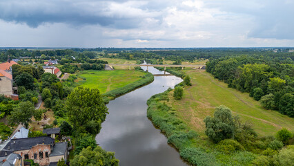 Fototapeta premium Aerial panorama of the Warta River in the town of Pyzdry. Green riverbanks, a bridge, and surrounding buildings create a peaceful rural landscape.