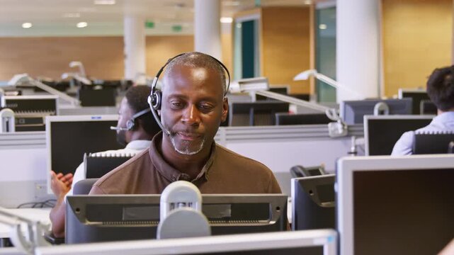 Camera moves around casually dressed mature businessman wearing telephone headset, working in customer service call centre - shot in slow motion