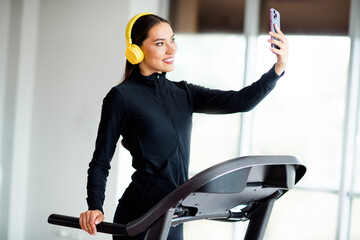 Athletic woman taking a selfie on a treadmill while wearing yellow headphones in a bright modern...