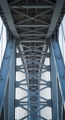 A dynamic shot from directly underneath a large, modern bridge's steel girders, emphasizing the repeating geometric patterns and imposing scale. The light filters through the structure, casting intere
