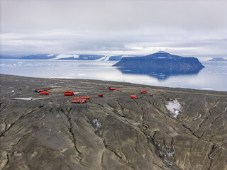 Aerial view of red buildings contrasting against the stark, dark landscape with a backdrop of icy water and a distant island, Seymour Island, Antarctica.