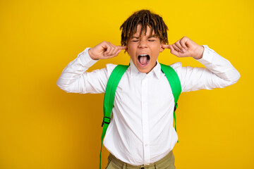 Young schoolboy shouting with fingers in ears against yellow background expressing frustration in...