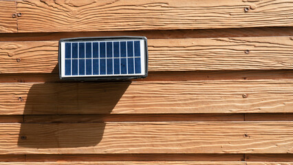 Small solar panel is mounted on wooden wall, casting shadow in sunlight. wood has textured, natural...