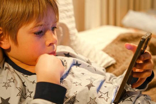 Cute kid boy in pajamas lying in bed watching smartphone, smiling. Children's screen time. Child plays games using phone at the evening or night. Social media addiction. Childhood, modern technology