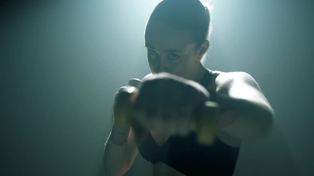Młoda blondynka boksuje w stronę kamery w ciemnej sali treningowej. W rękach trzyma ż&oacute;łte ciężarki, światło zza niej tworzy poświatę, a jej skupiona twarz dodaje dramatyzmu scenie