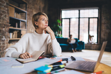 Freelancer woman reflects on interface concept while resting her chin on hand, holding notebook,...