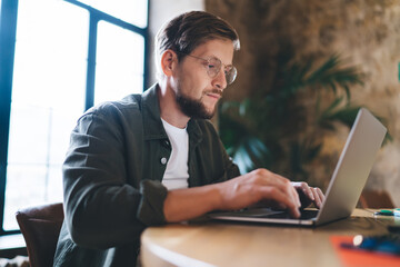 Side view of bearded male freelancer working with concentration, typing on laptop, immersed in creative flow and digital execution at round wooden table.