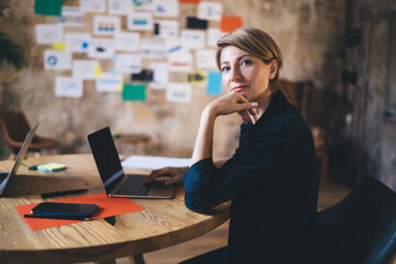 Smart female freelancer gazing at viewer while typing on laptop, showing clarity, confidence and digital independence in bright, tech-friendly creative office space