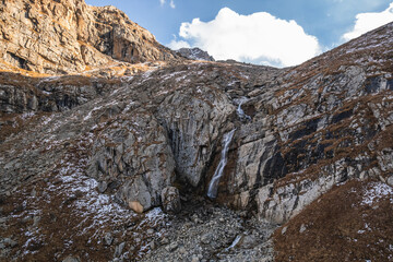 Serene waterfall cascading down rugged mountainside in the Caucasus Mountains.  Sunlight illuminates snow-dusted rocks and dry grass. A stunning natural landscape.