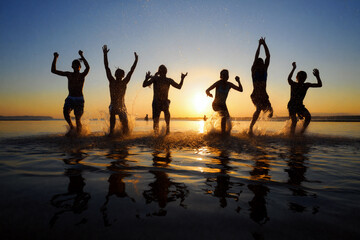 Obraz premium A group of six friends joyfully splashes in the water at a beach, silhouetted against a vibrant sunset, creating a lively atmosphere of fun and friendship. summer holidays concept. Generative AI.