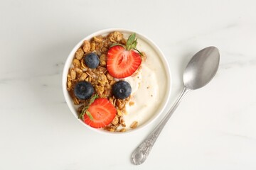Tasty yogurt with granola, berries and spoon on white marble table, flat lay