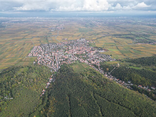 Scenic aerial view of a picturesque European village with red-tiled roofs nestled between lush green forests and patchwork farmlands under a moody overcast sky, ideal stock photo.