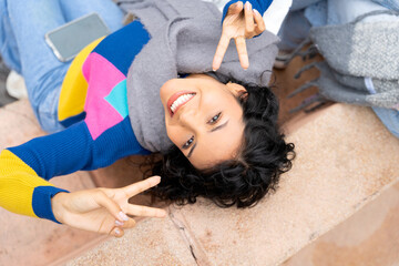 Young latin american woman gesturing peace sign lying on city steps