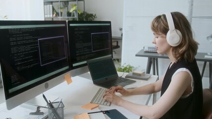 Young adult woman programming while listening to music via headphones. She sitting at wooden desk surrounded by two huge monitors and laptops - Powered by Adobe
