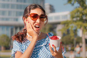 Young woman is holding a cup of ice cream and experiencing a sudden toothache due to its coldness,...
