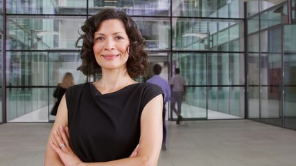 Mature businesswoman with shoulder bag standing in busy foyer of modern office building with timelapse of colleagues leaving and arriving in background 