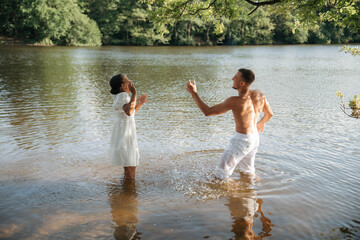 Romantic Interracial Couple Dancing Barefoot on Beach by the Lake, Wearing White Clothes, Smiling and Enjoying Summer Day Outdoors, Love and Togetherness in Nature