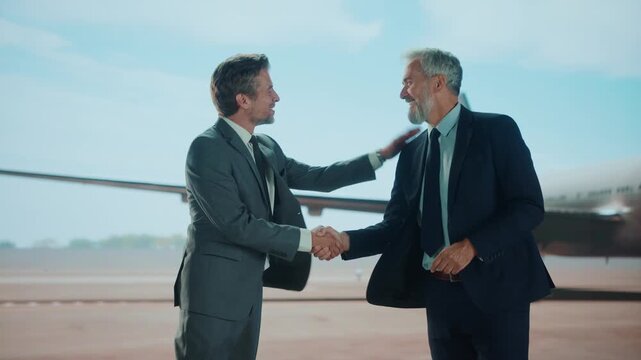 Two businessmen in suits shaking hands on the tarmac with an airplane in the background, possibly after a successful business trip or meeting.