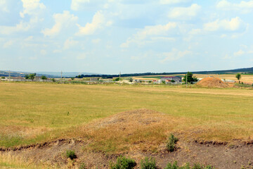Obraz premium A vast open field with dry grass and scattered shrubs under a blue sky with fluffy clouds. The landscape features distant hills and a few buildings