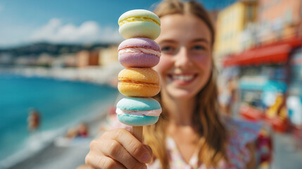 Woman smiling while holding stacked, colorful macarons, with a beautiful beach and Mediterranean sea creating a picturesque background