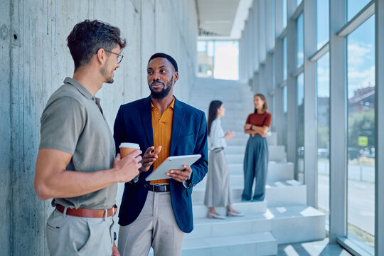 Two businessmen discussing work while holding tablet and drinking coffee, with colleagues talking in the background