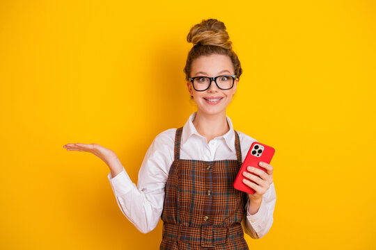 Smiling teenage student holding a smartphone and gesturing with her hand against a vibrant yellow background