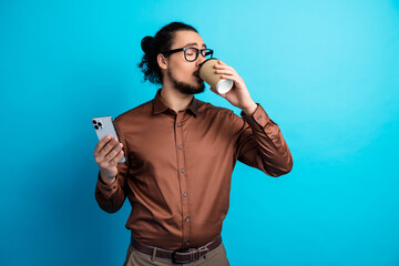 Young man in brown formal shirt holding smartphone and drinking coffee on turquoise background...