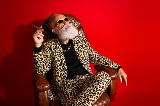 Eccentric elderly gentleman in vibrant leopard suit with sunglass seated against vivid red background representing style and charisma