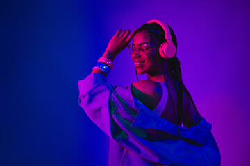 Young woman enjoying music with stylish headphones under vibrant neon lighting, showcasing fashion...