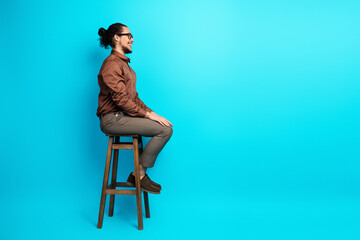 Portrait of a young businessman seated on a high stool against a light turquoise background,...