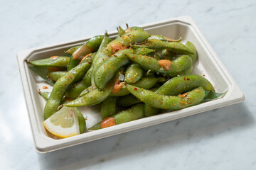 Fresh edamame with hot sauce in a takeout container on a white background. Natural light enhances this healthy Japanese dish, perfect for Asian fusion cuisine lovers