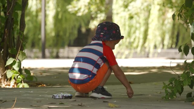 A young boy draws with chalk on a paved path, surrounded by lush greenery. He enjoys a quiet and sunny moment of creative play in a park in Simferopol, Crimea.