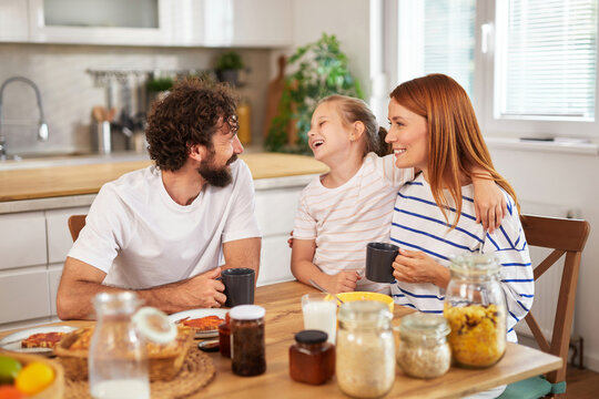 A family sits around the dining table, enjoying breakfast. The parents share smiles while their child laughs, creating a warm and joyful atmosphere in the bright kitchen.