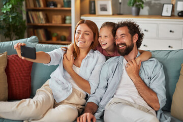 A family of three, including a mother, father, and young daughter, smiles and poses for a selfie on a comfortable couch filled with pillows. They appear happy and relaxed.