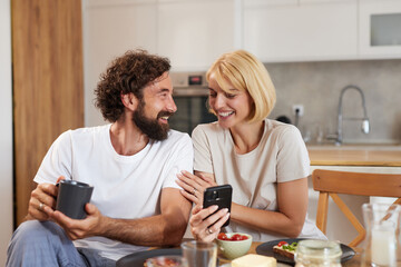 A couple shares a light-hearted moment at a table in a modern kitchen, smiling at a smartphone,...