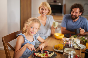 A family gathers around the breakfast table, smiling and sharing moments. The daughter playfully...