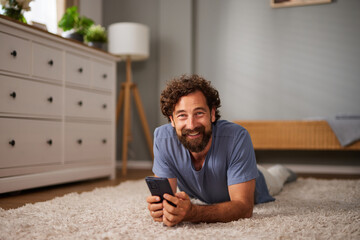A relaxed man lies on a plush carpet, smiling as he engages with his smartphone in a warm and...